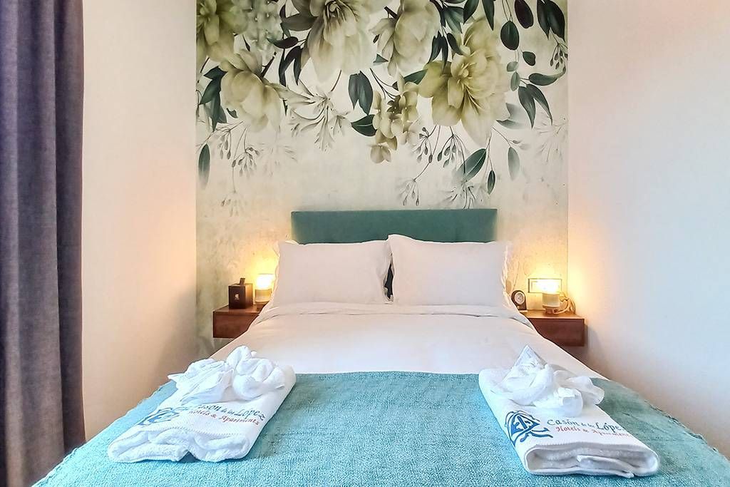 Dormitorio con papel tapiz floral, colcha verde azulado y dos mesitas de noche con lámparas.
