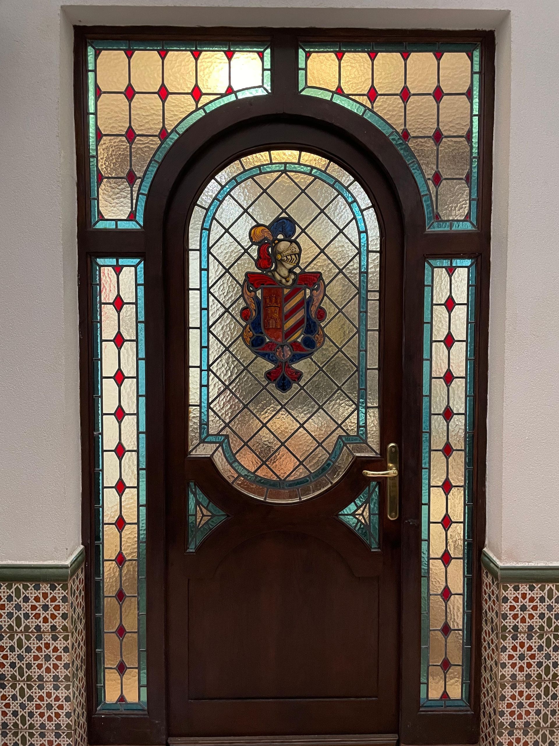Puerta de madera oscura con paneles de vidrio de colores y escudo.
