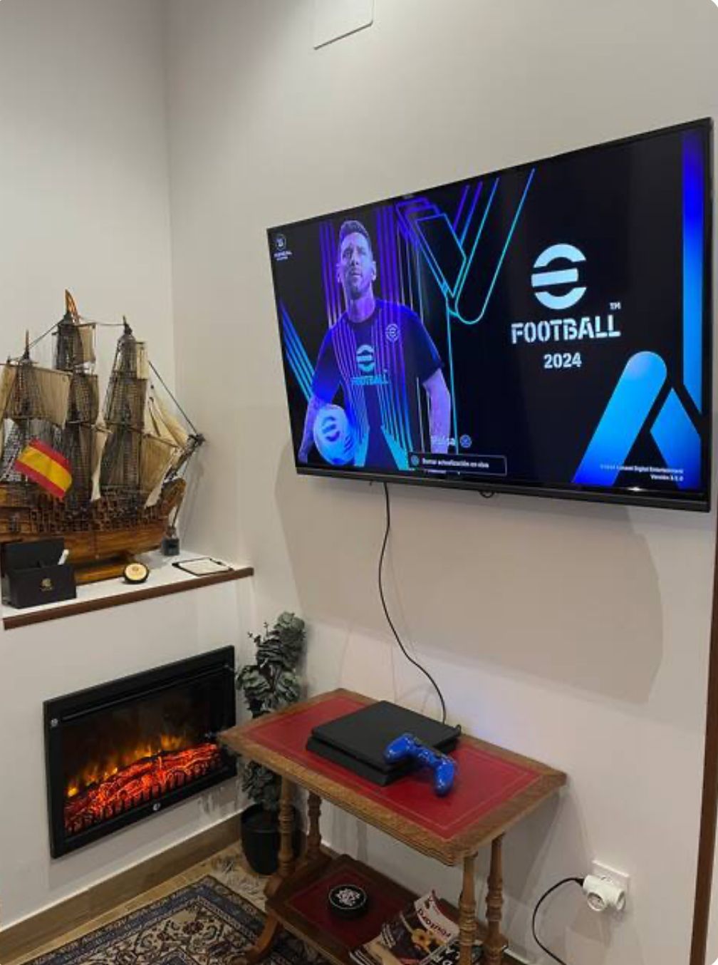 Configuración de juego: TV con juego de fútbol, ​​consola, mesa pequeña con control, chimenea, maqueta de barco en un rincón de una habitación.