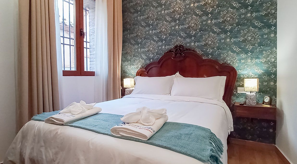 Acogedor dormitorio con cama doble, sábanas blancas, manta turquesa y cabecero de madera. Una ventana a la izquierda y dos lámparas de noche.