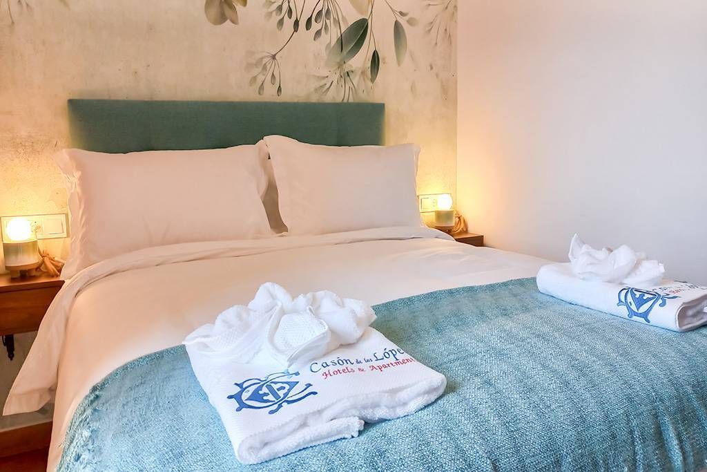 Acogedor dormitorio con una cama doble, detalles en verde azulado, ropa de cama blanca y papel tapiz floral.