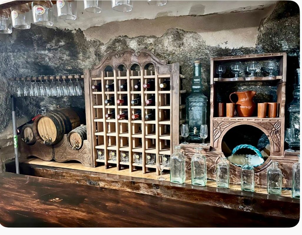 Un bar rústico con botellas, vasos y estantes de madera. Barriles y una estructura decorativa a la izquierda.