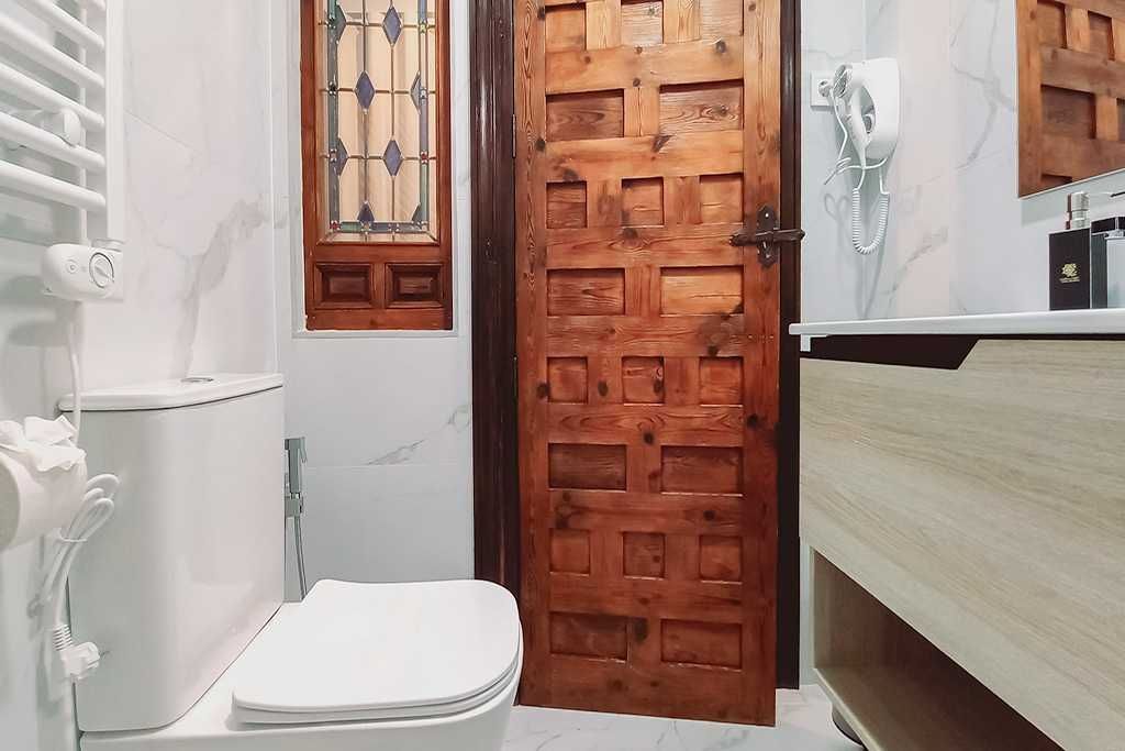 Baño con puerta de madera, inodoro y lavabo. Tonos blancos y marrones, paredes de mármol.