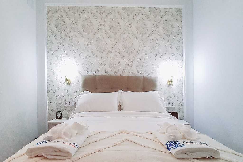 Dormitorio con cama doble, papel pintado estampado, sábanas blancas y lámparas de mesa.