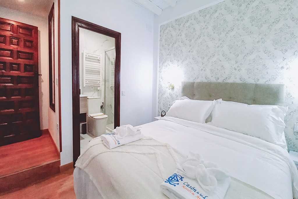 Dormitorio con cama, puerta de baño y puerta de madera. Ropa de cama blanca y papel pintado estampado.