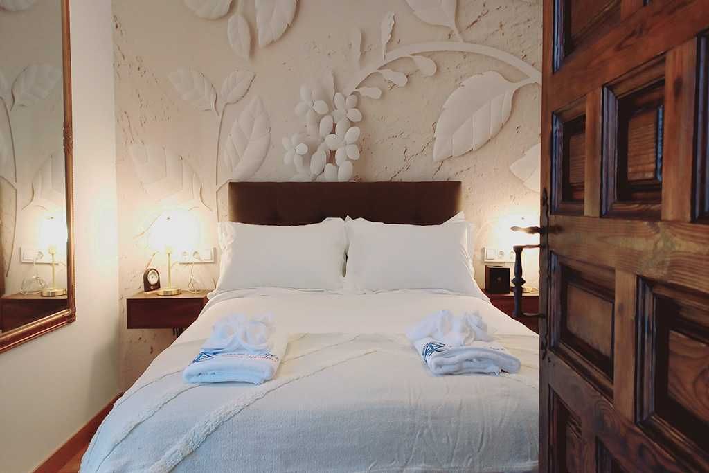 Interior del dormitorio: ropa de cama blanca, pared floral, puerta de madera abierta.
