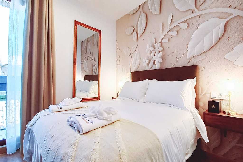 Dormitorio con ropa de cama blanca, arte floral en la pared y un espejo con marco de madera.