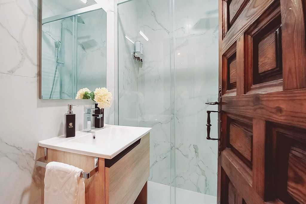 Baño con paredes de mármol, ducha de cristal, tocador de madera y puerta de madera abierta.