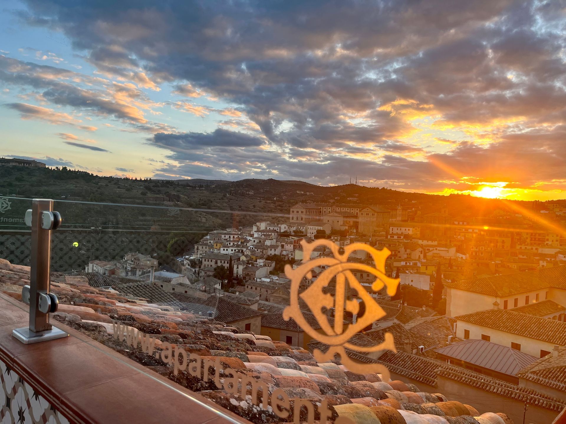 Atardecer sobre un paisaje urbano con un emblema metálico en un tejado. Cálidos tonos naranjas y amarillos en el cielo.