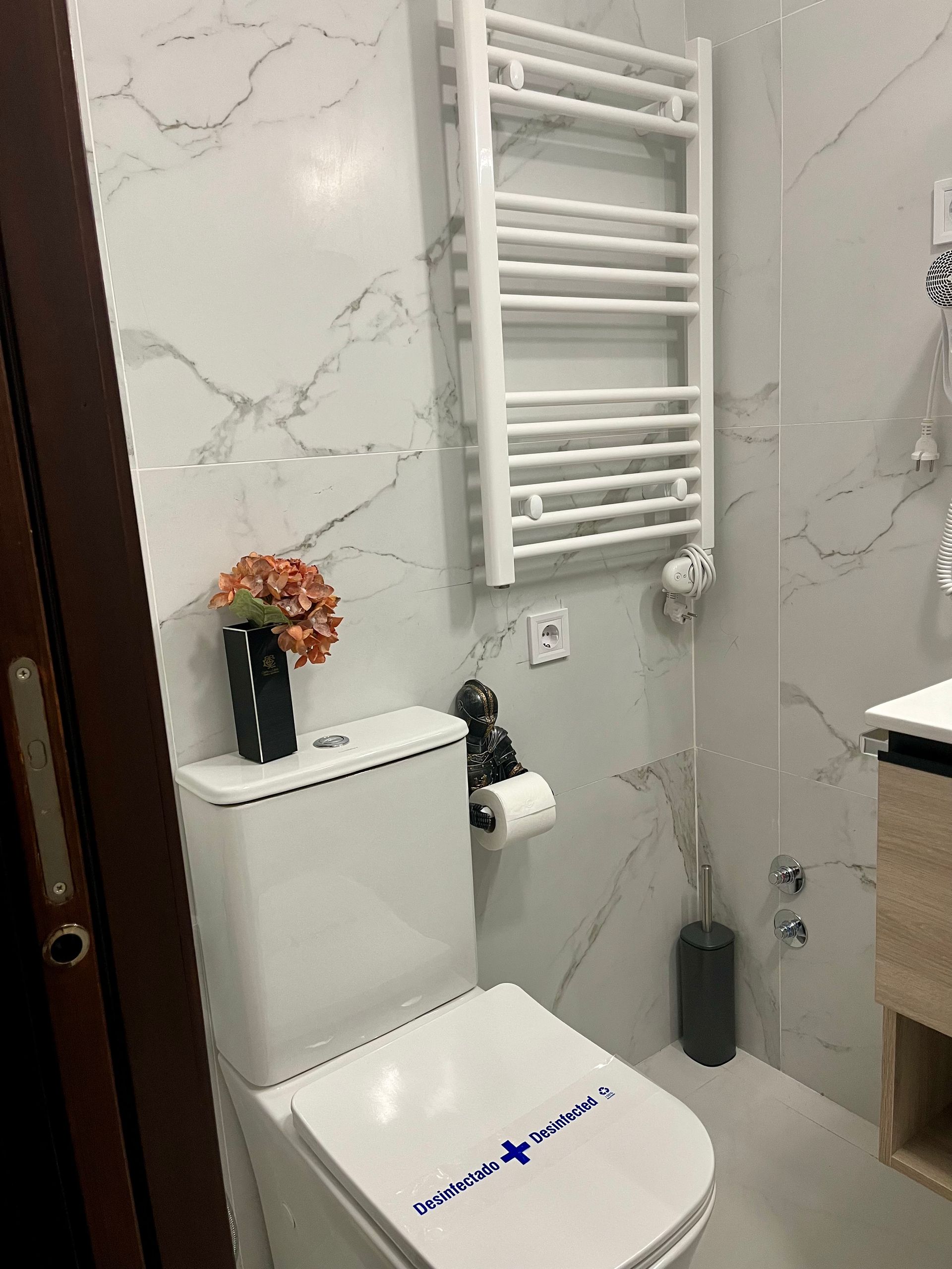 Baño blanco con inodoro, toallero y arreglo floral sobre baldosas con estampado de mármol.