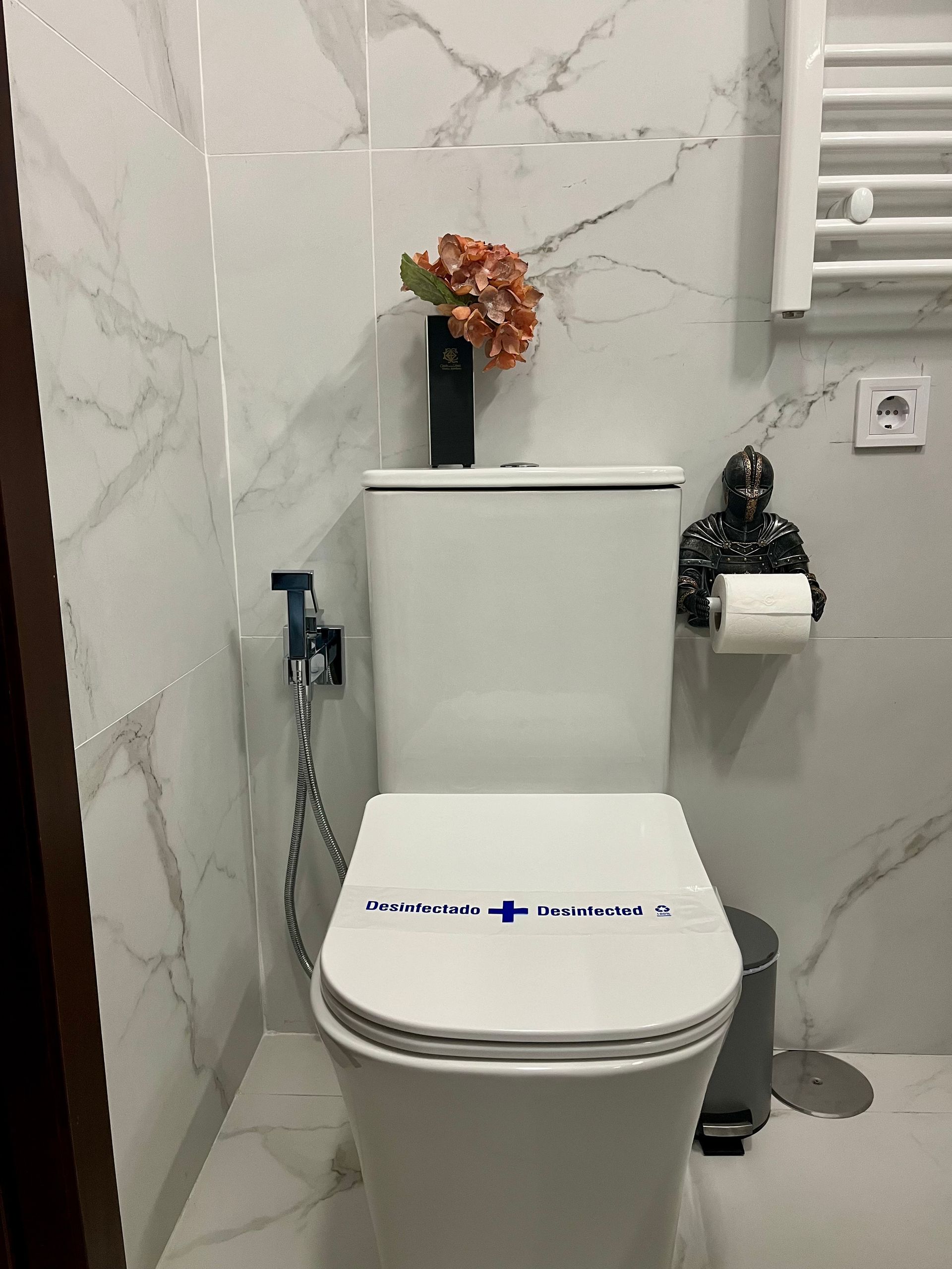 Inodoro blanco con arreglo floral, bidé y portarrollos de papel higiénico decorativo contra una pared con diseño de mármol.