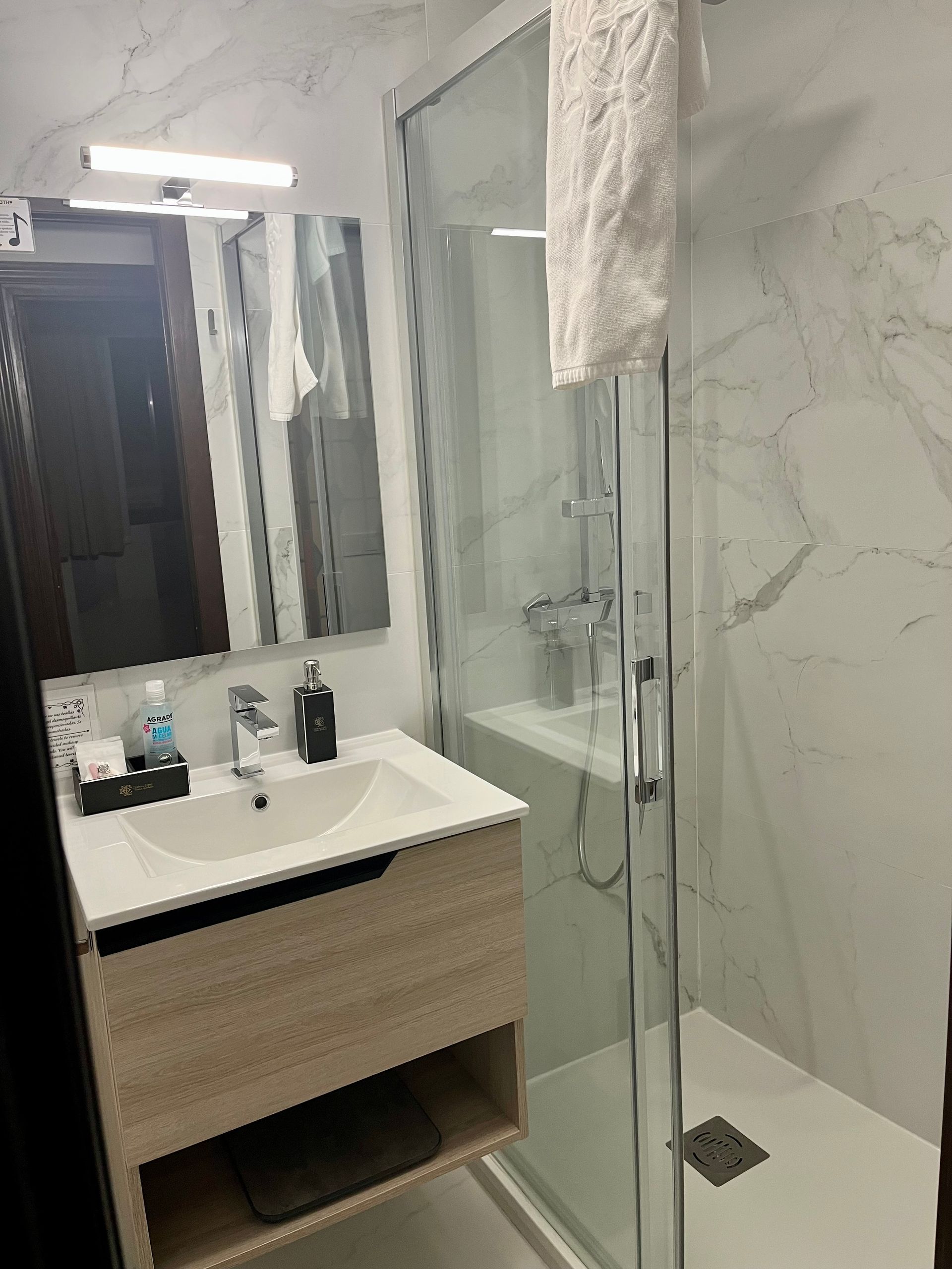 Pequeño baño moderno con ducha, lavabo y pared con efecto mármol. Hay una toalla blanca colgada.