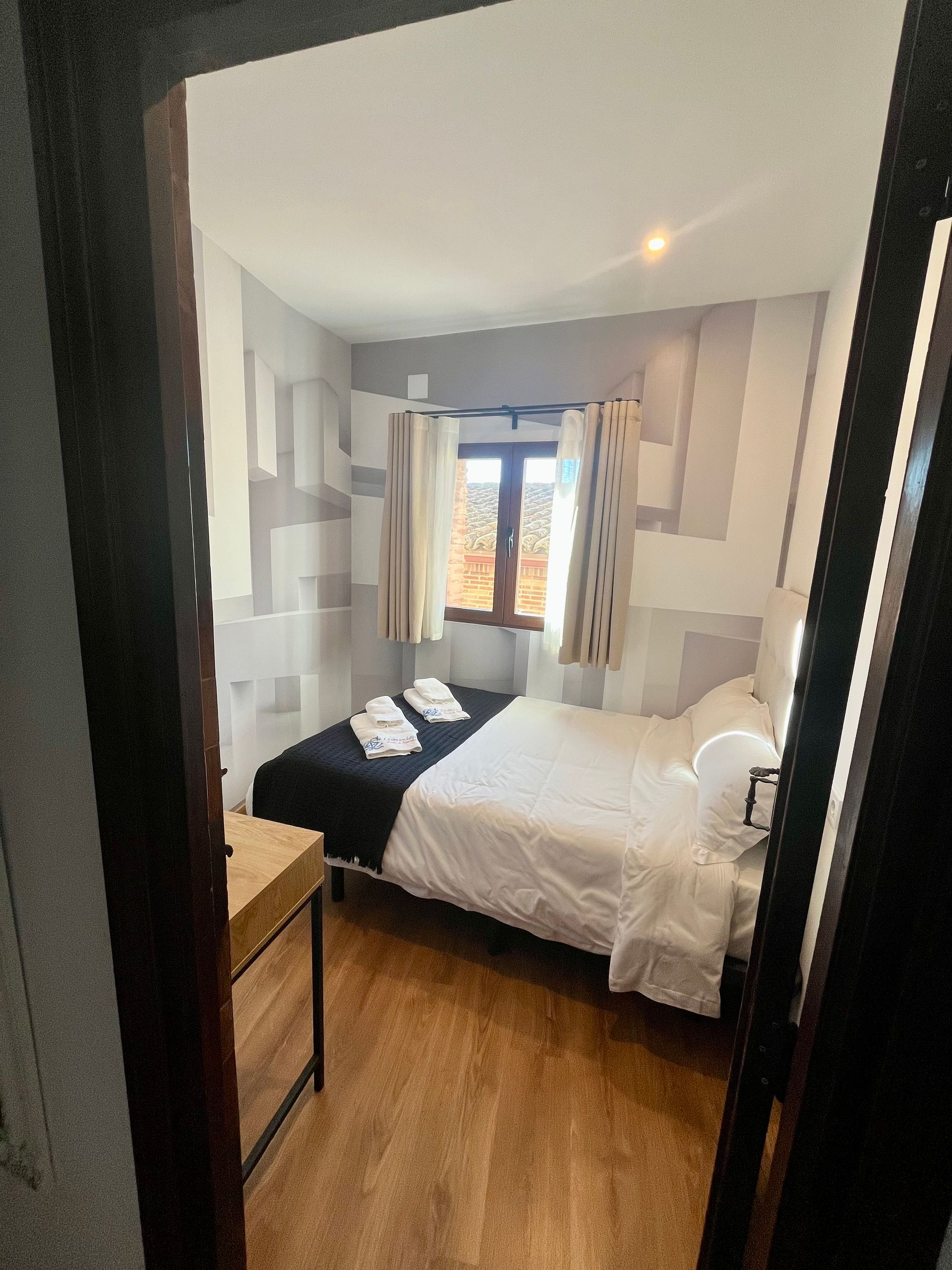 Dormitorio con cama, ventana y suelo de madera, visible a través de una puerta abierta. Las paredes presentan un estampado blanco y gris.