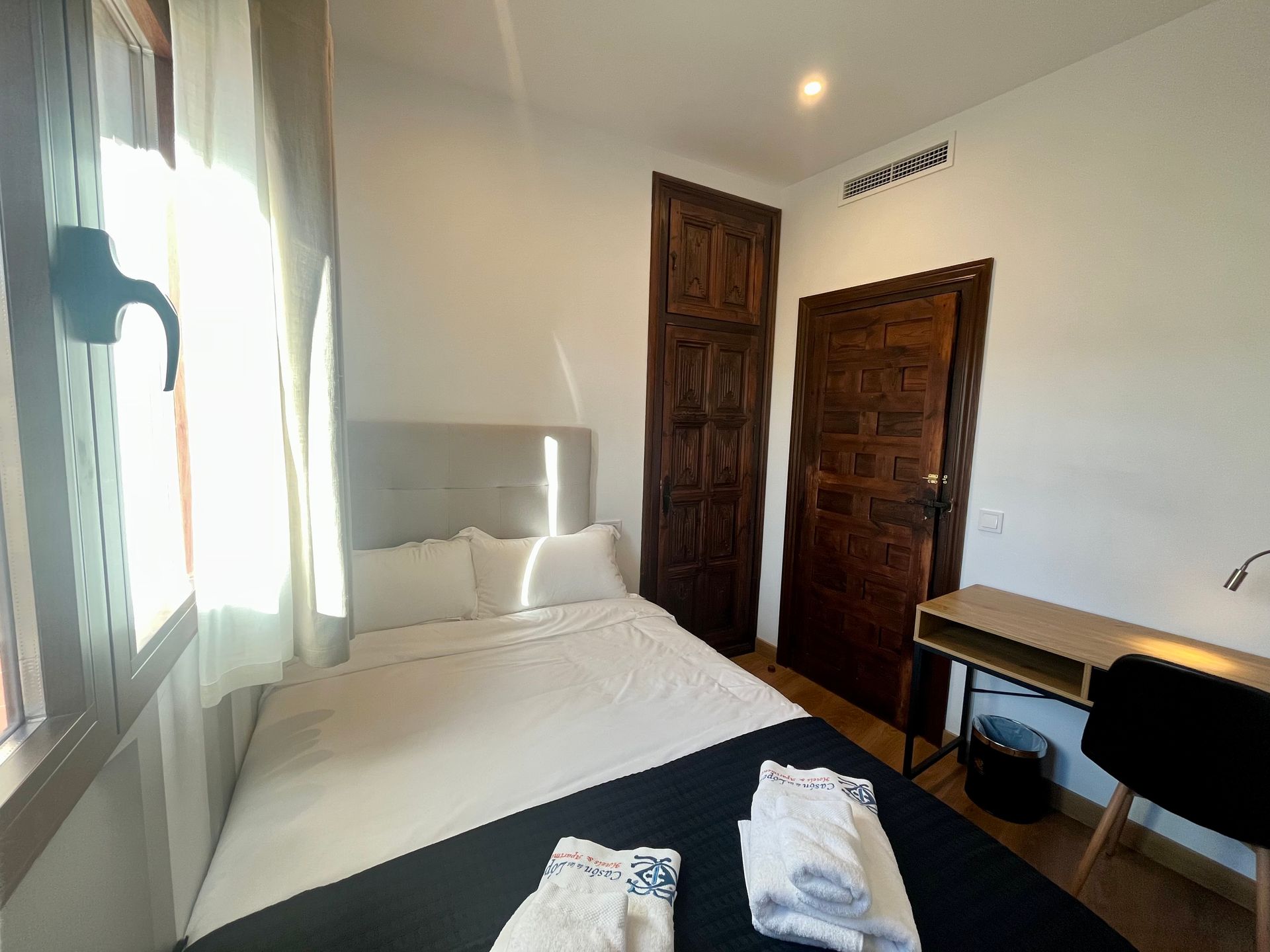 Dormitorio con una cama, dos puertas marrones, un escritorio y una ventana por donde entra la luz del sol.