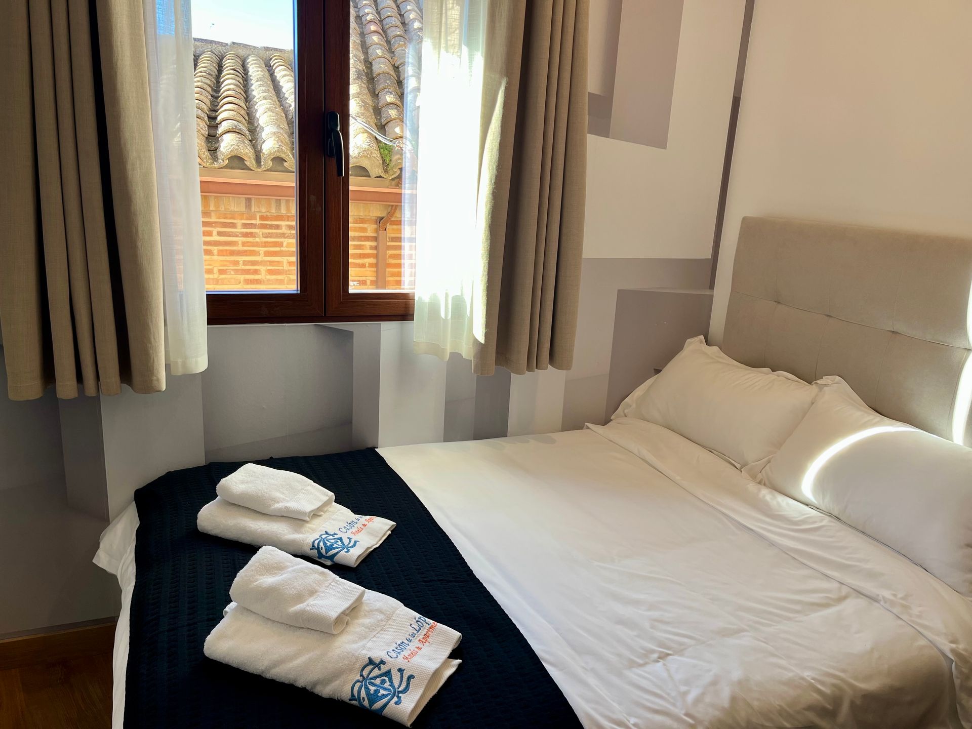Dormitorio con cama blanca, ventana y toallas. Paredes grises y blancas, con un atisbo de tejado.