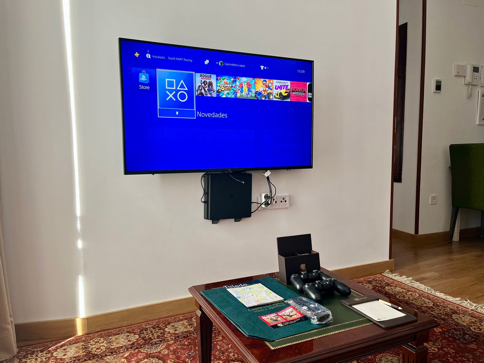 Un televisor montado en la pared con una configuración de PlayStation 4. Juegos mostrados en pantalla, controlador y mesa debajo.