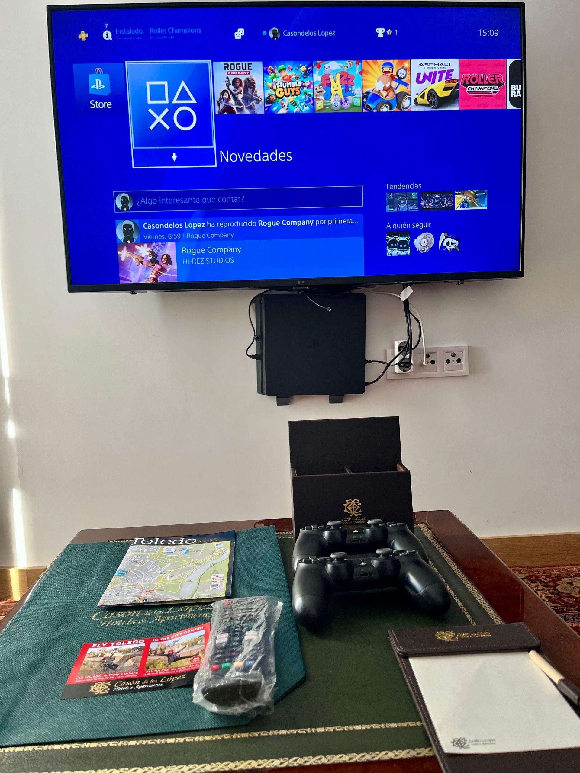 Configuración de PlayStation 4 sobre una mesa con controlador y control remoto. TV montada en la pared, consola debajo.