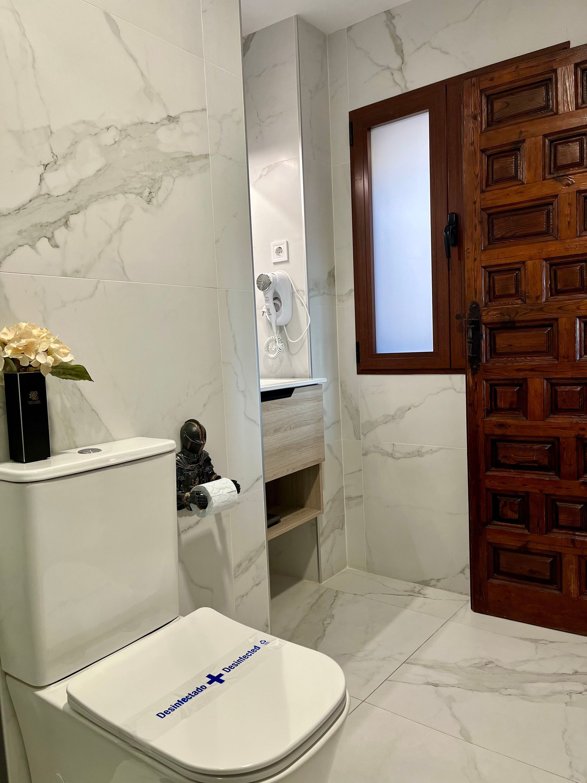 Baño con paredes de mármol blanco, inodoro, puerta de madera y ventana esmerilada.