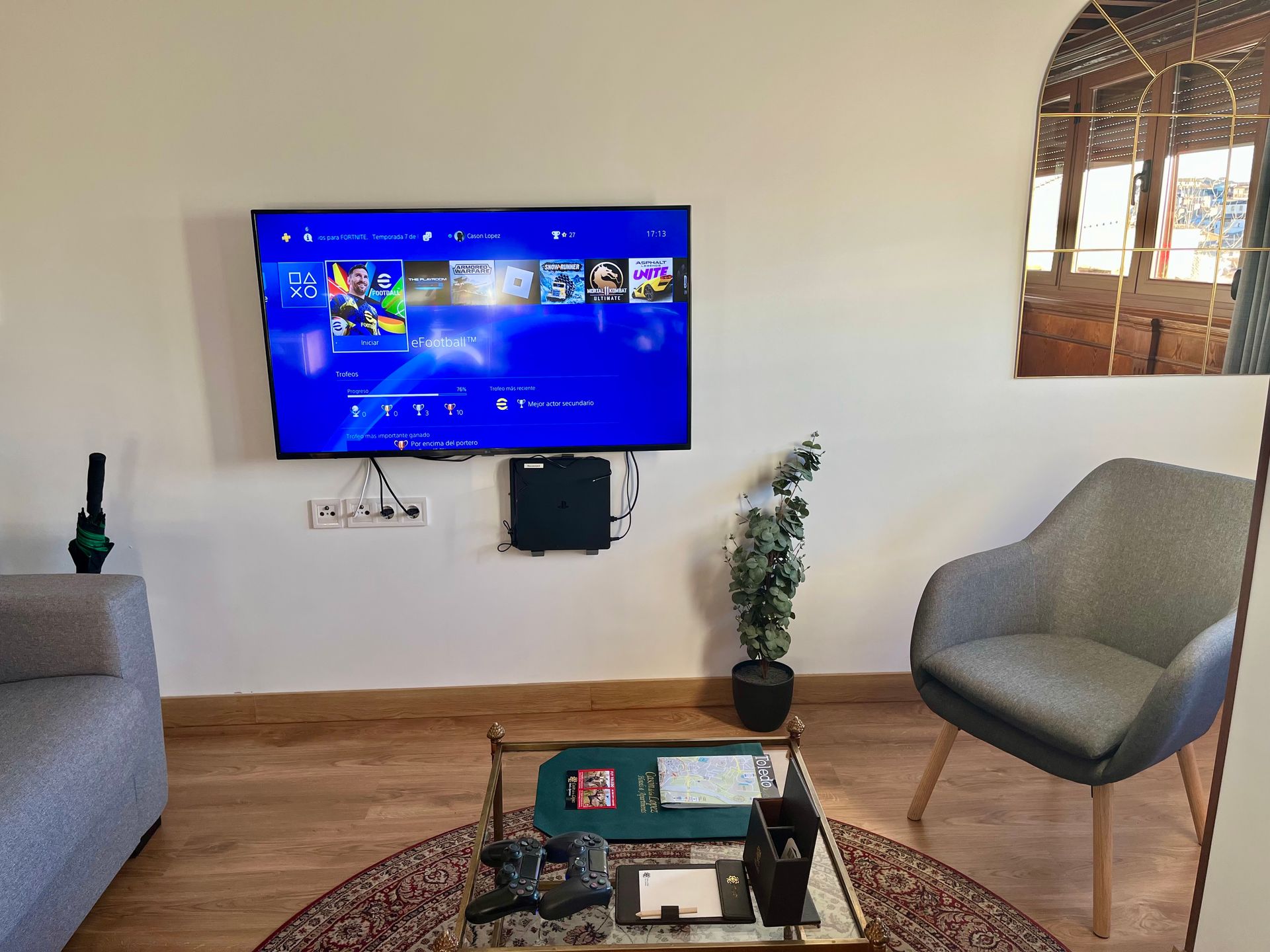 Una sala de estar con un televisor montado, una PS4, un sillón gris y una mesa de café.