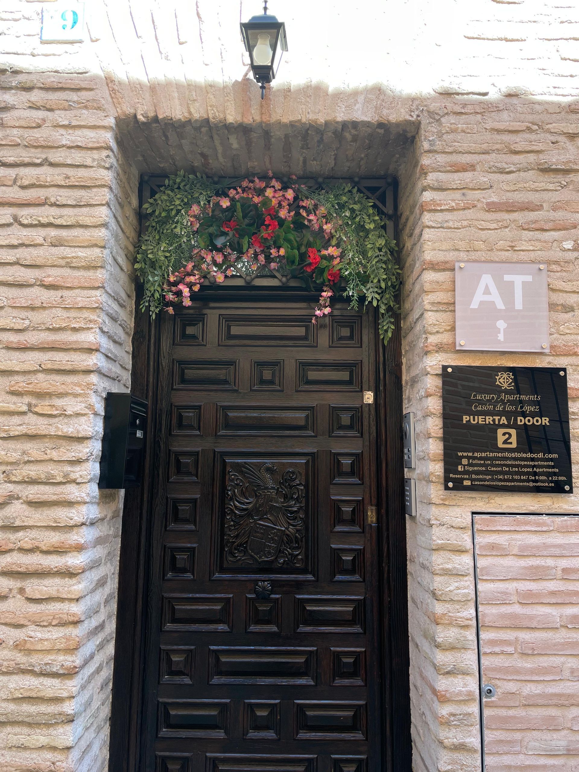 Puerta de madera oscura con guirnalda floral, situada en un arco de ladrillo, junto a una placa que dice 