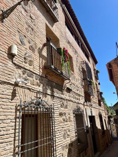 Edificio de ladrillo con balcones, rejas en las ventanas y plantas con flores; en una calle estrecha bajo un cielo azul.