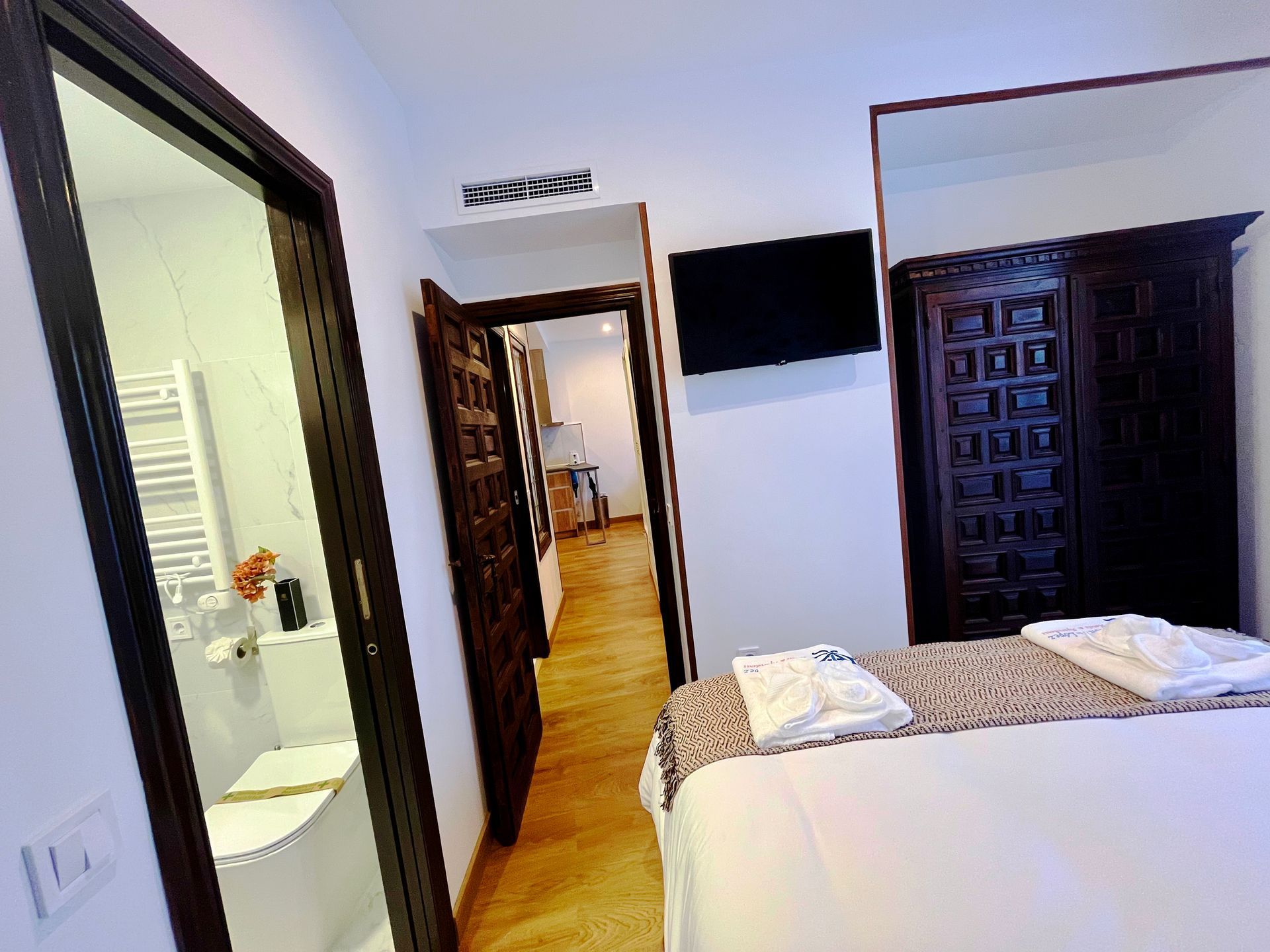 Dormitorio con cama, armario oscuro, puerta del baño a la izquierda, TV en la pared.