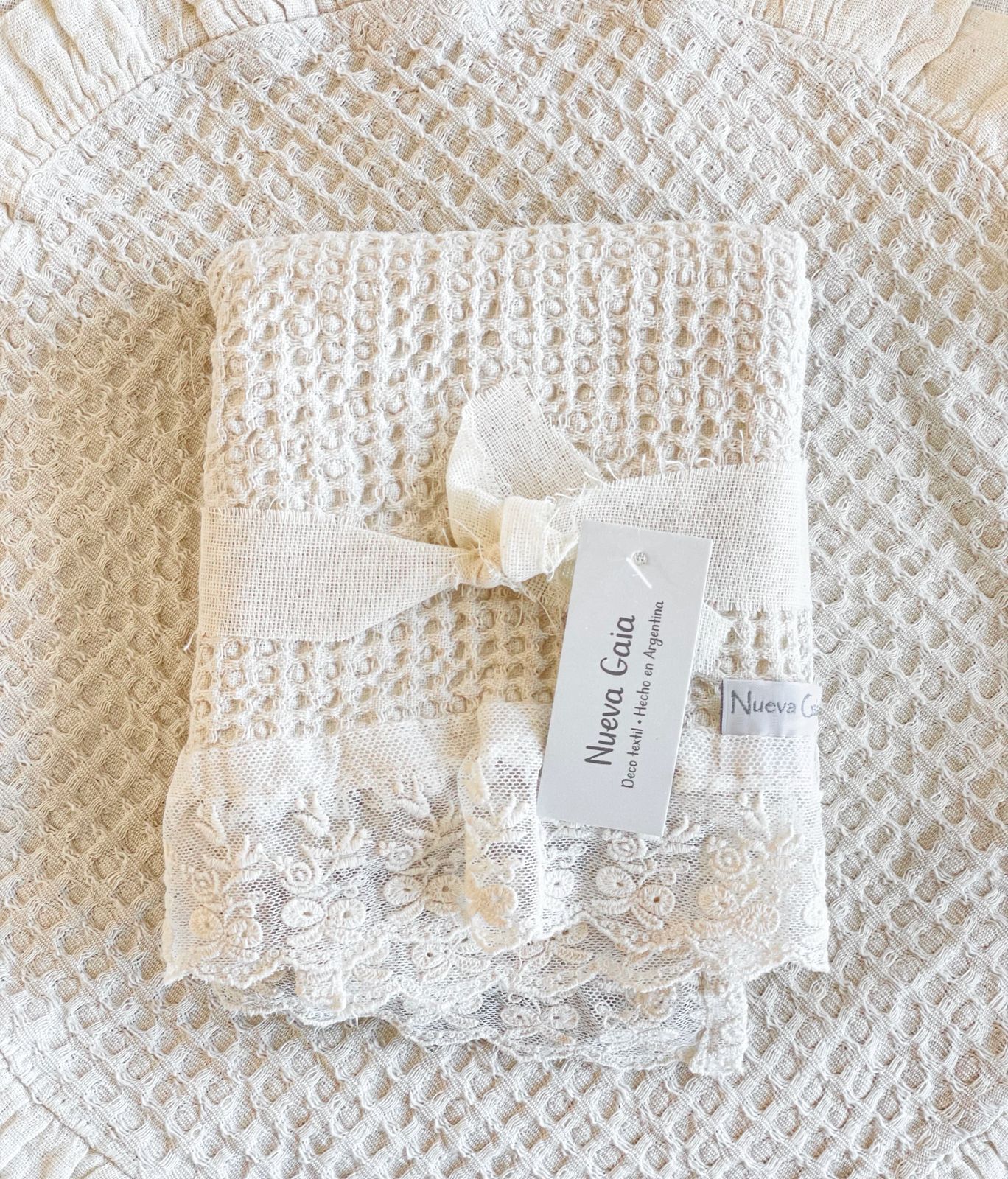 Manta blanca de encaje para bebé con un pequeño lazo de cinta y etiqueta sobre un fondo acolchado texturizado color crema.