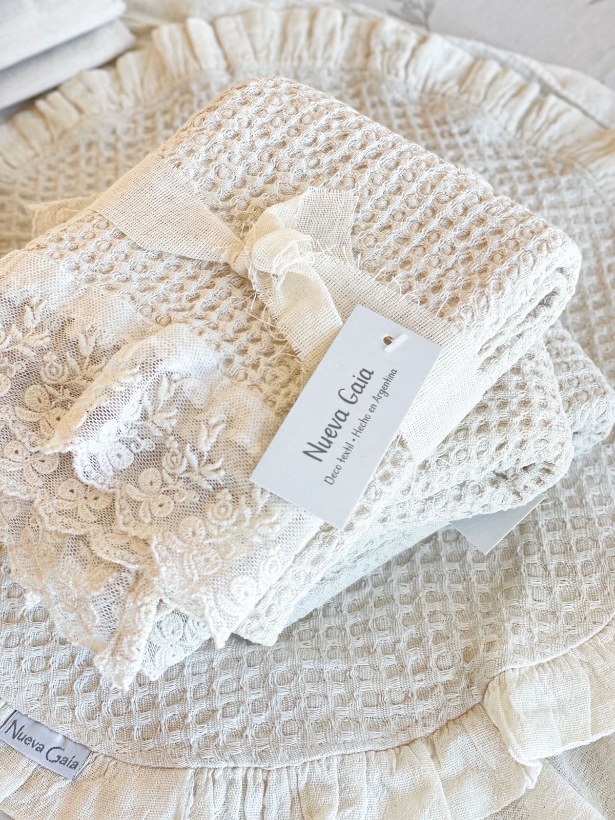 Manta blanca de encaje para bebé con un pequeño lazo de cinta y etiqueta sobre un fondo acolchado texturizado color crema.