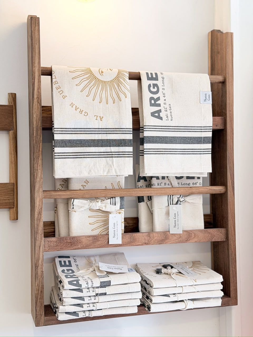 Estantería de madera tipo escalera que sostiene textiles doblados y revistas en una habitación de tonos neutros.