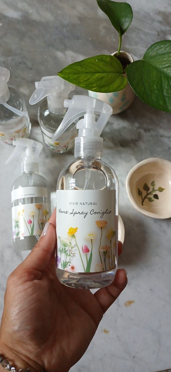 Una mano sostiene un frasco de spray floral con dos frascos iguales sobre una superficie de mármol y una planta verde cerca.
