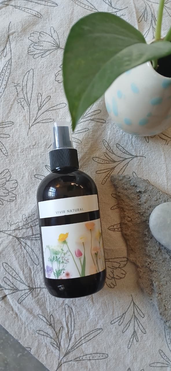 Una mano sostiene un frasco de spray floral con dos frascos iguales sobre una superficie de mármol y una planta verde cerca.