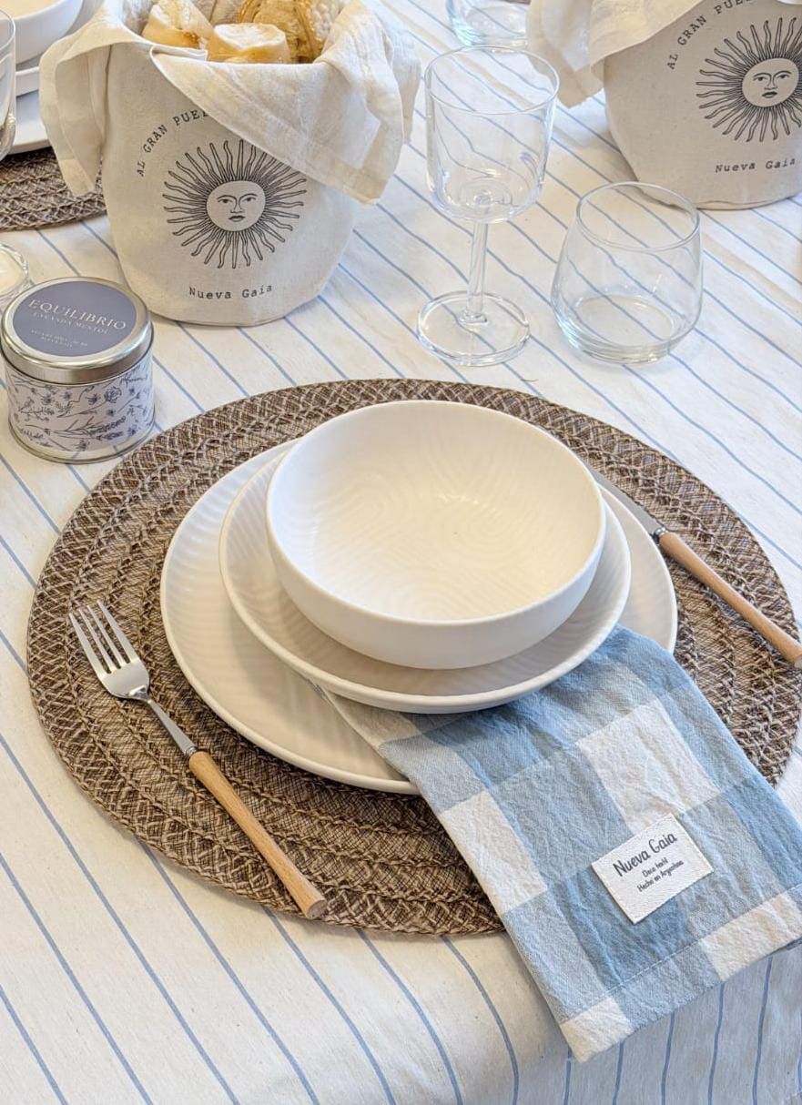 Mesa festiva con vajilla blanca, manteles individuales tejidos, servilletas de rayas azules y margaritas en un jarrón.