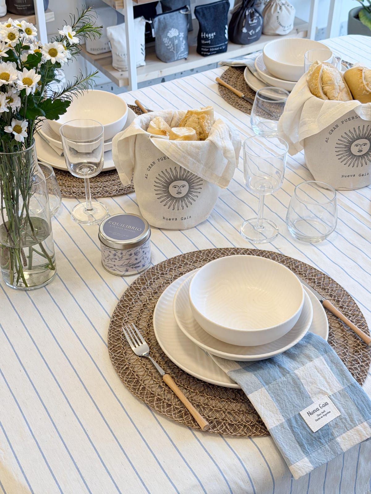 Mesa festiva con vajilla blanca, manteles individuales tejidos, servilletas de rayas azules y margaritas en un jarrón.