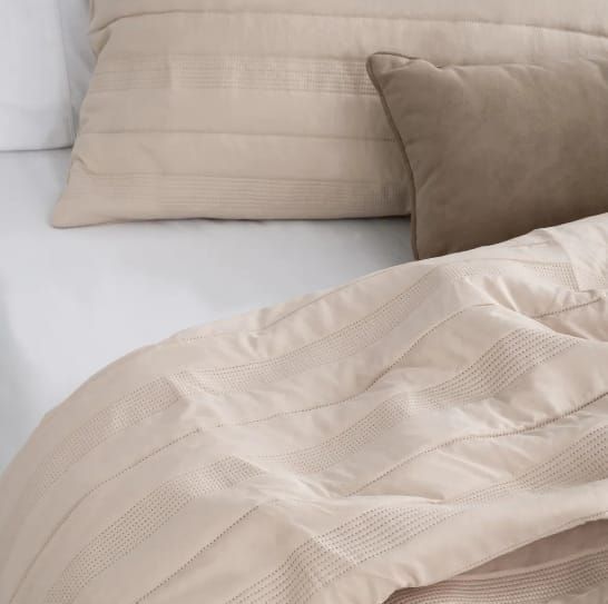Habitación luminosa de color beige con cama, mesita de noche, planta y rincón junto a la ventana de madera.