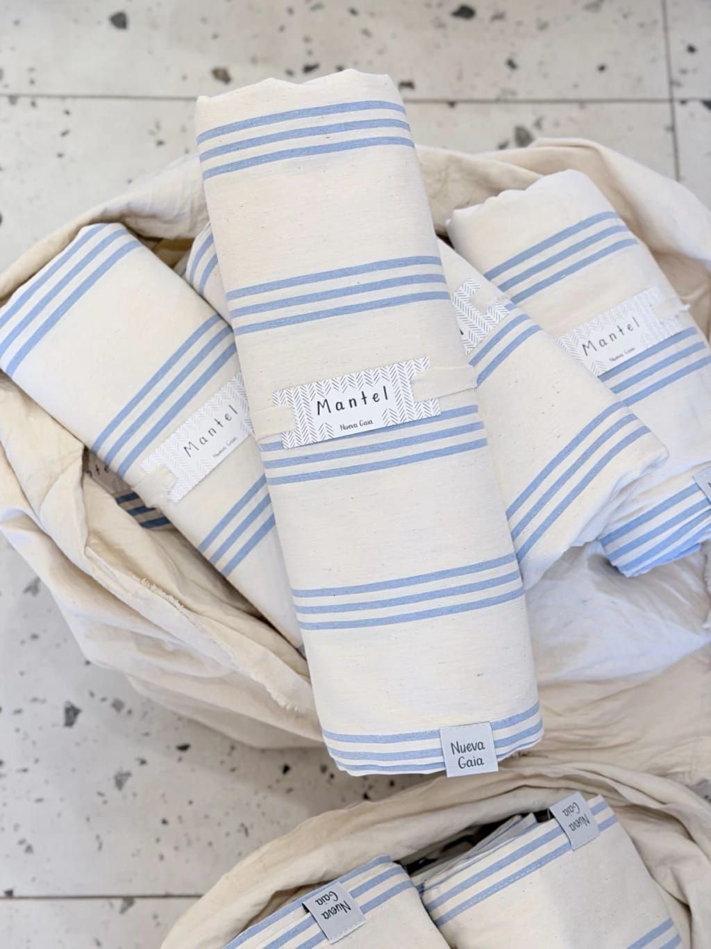 Tres paños blancos doblados con rayas azules en una bolsa de tela color crema sobre un suelo moteado.