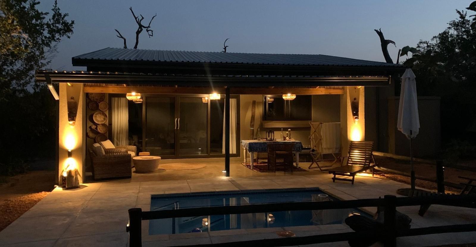 Night view of a lit patio with a pool, table, and furniture.
