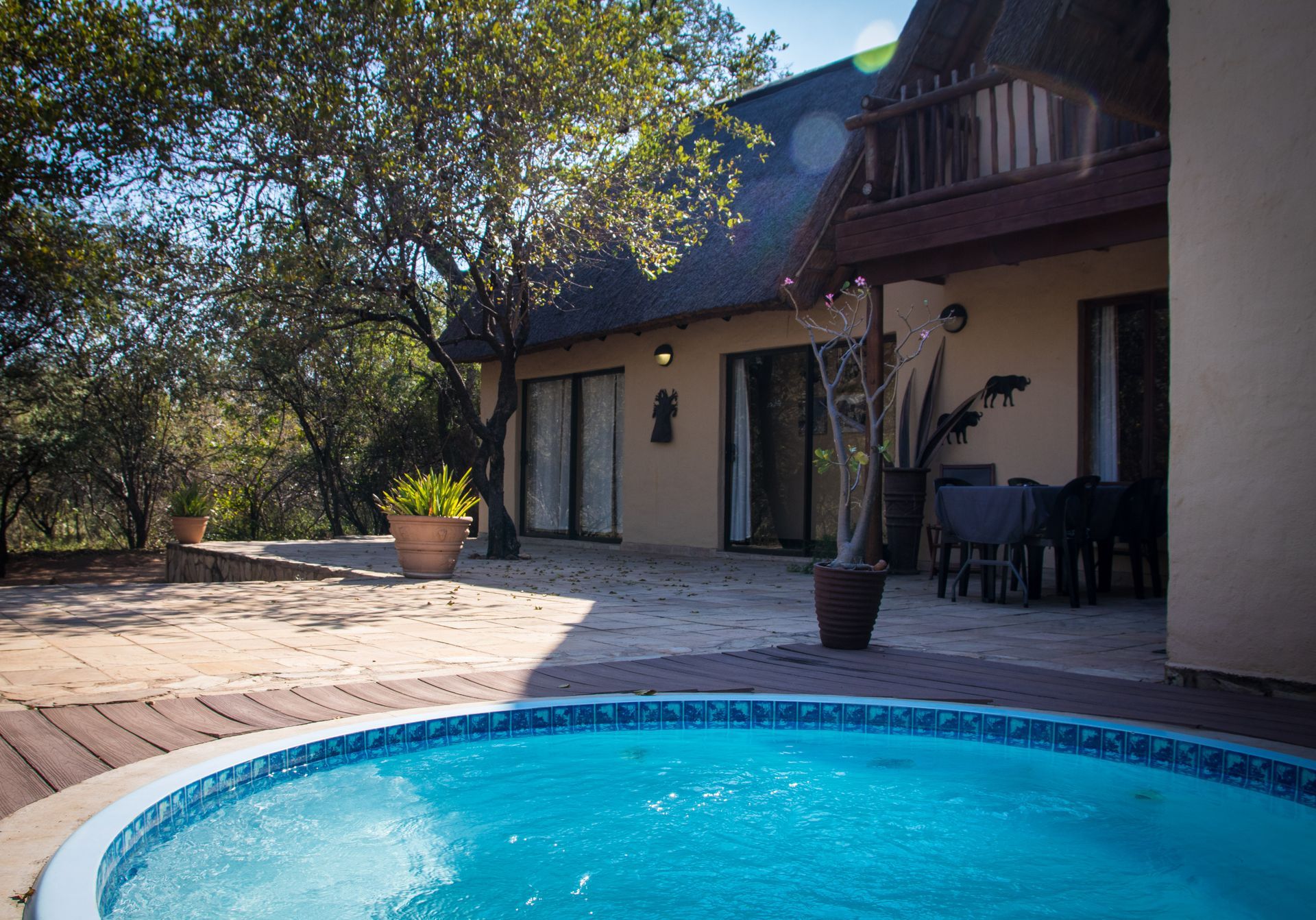 A pool with blue water, patio with table and chairs, and a thatch-roofed building in an outdoor setting.