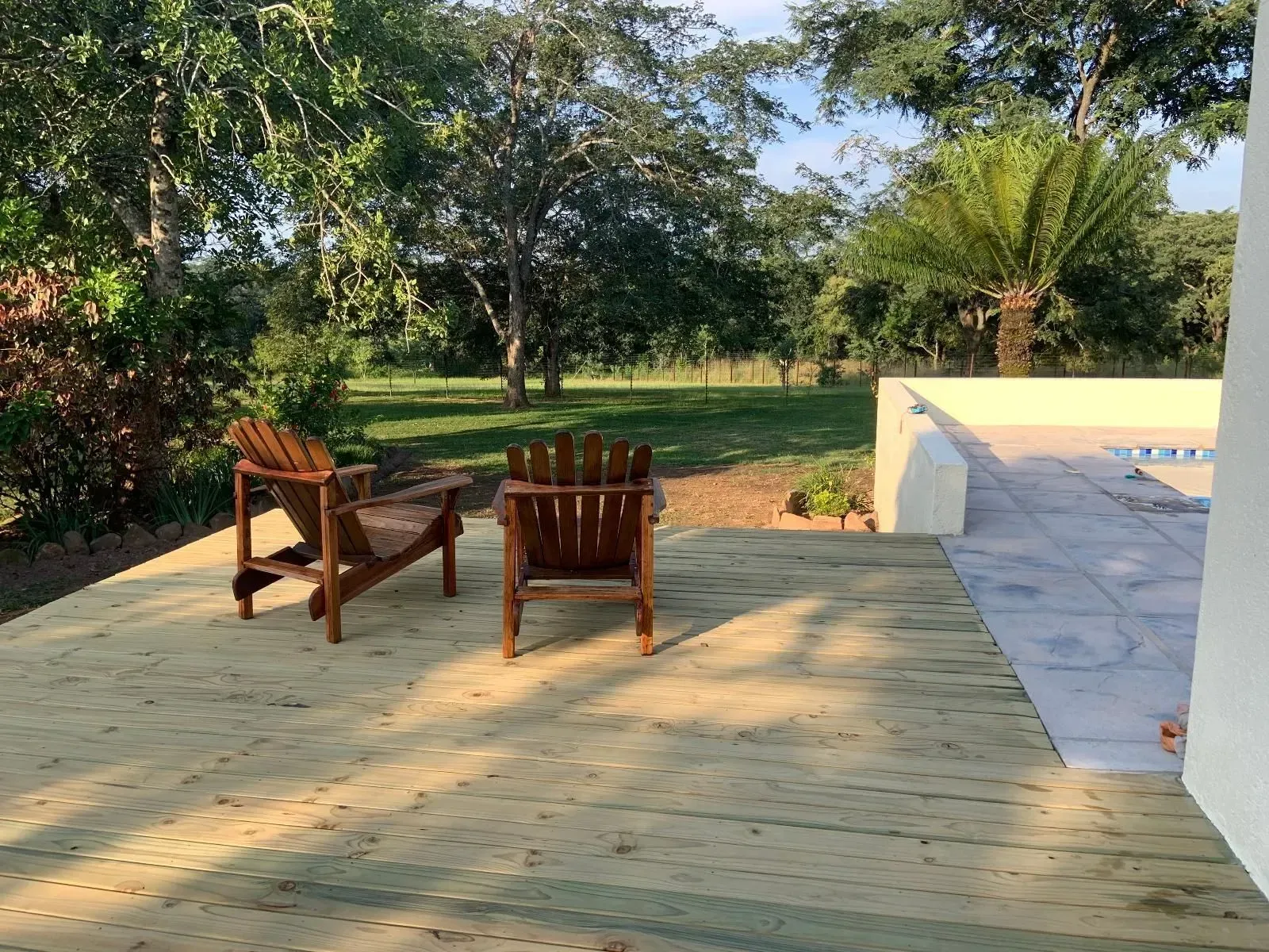 Two wooden Adirondack chairs on a sunlit wooden deck overlooking a yard and trees.