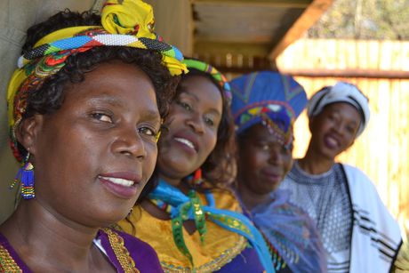 Four African women in colorful head wraps and dresses, smiling outdoors.
