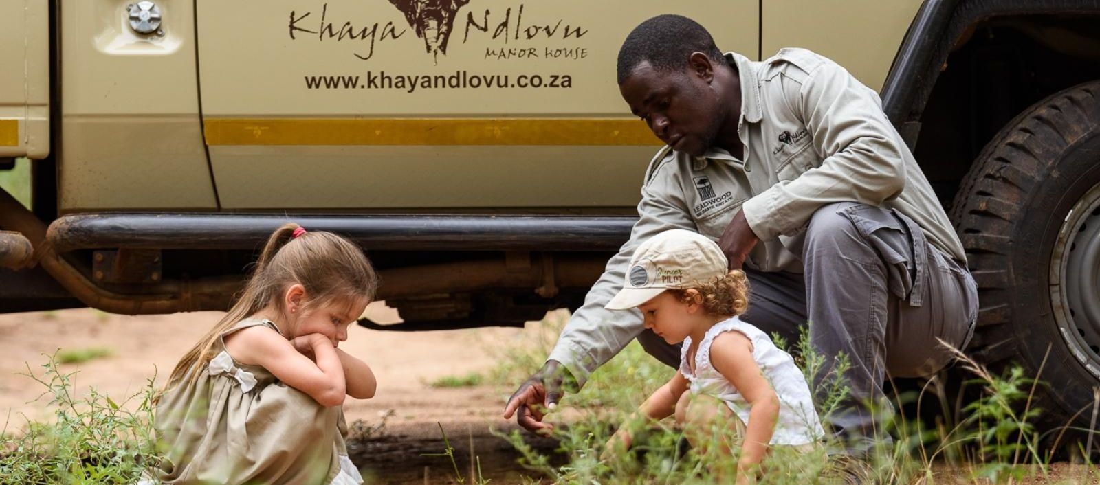 Family activities with Africana A man points at something in the grass for two children, near a safari vehicle.
