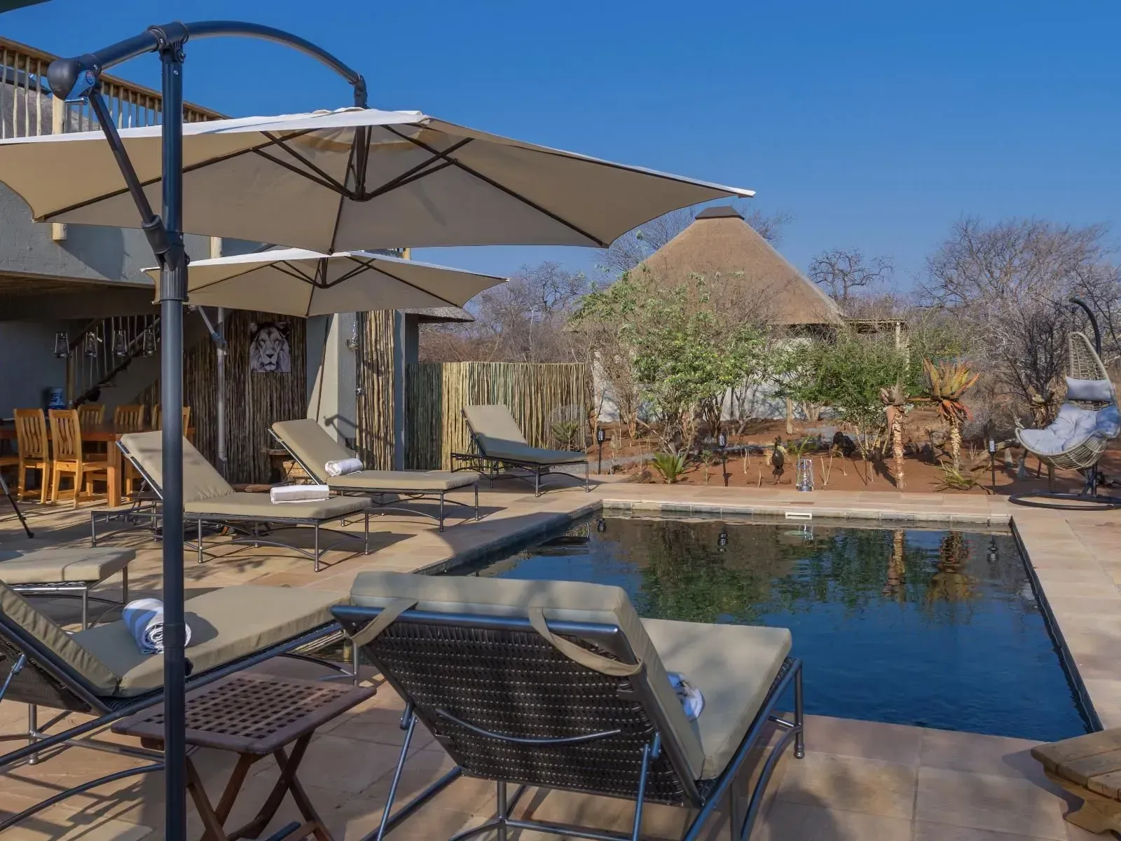 Hoedspruit Accommodation Poolside relaxation area with loungers, umbrellas, and a thatched-roof structure under a blue sky.