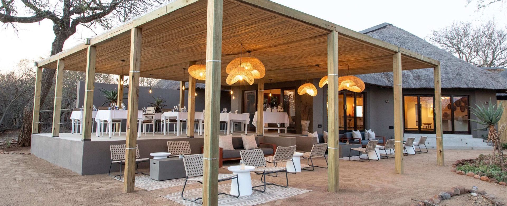 An open-air dining area with wooden structure, straw roof, tables, chairs, and hanging lights.