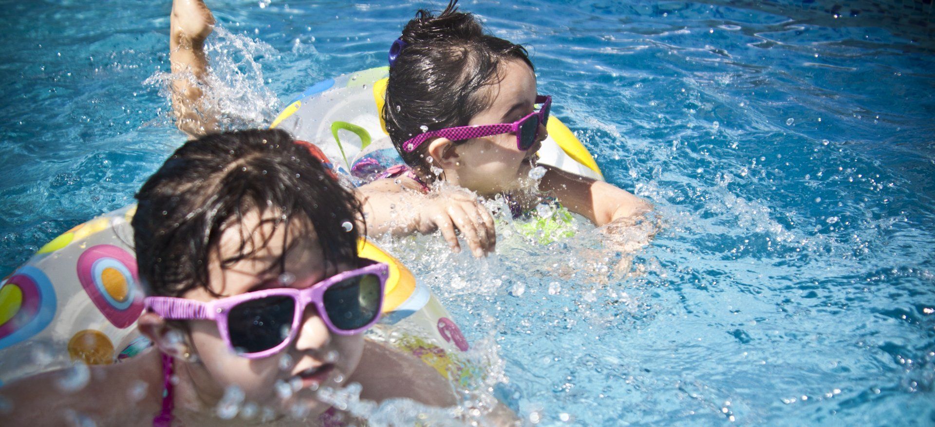 Two young girls in sunglasses swim in a pool, using inflatable rings.