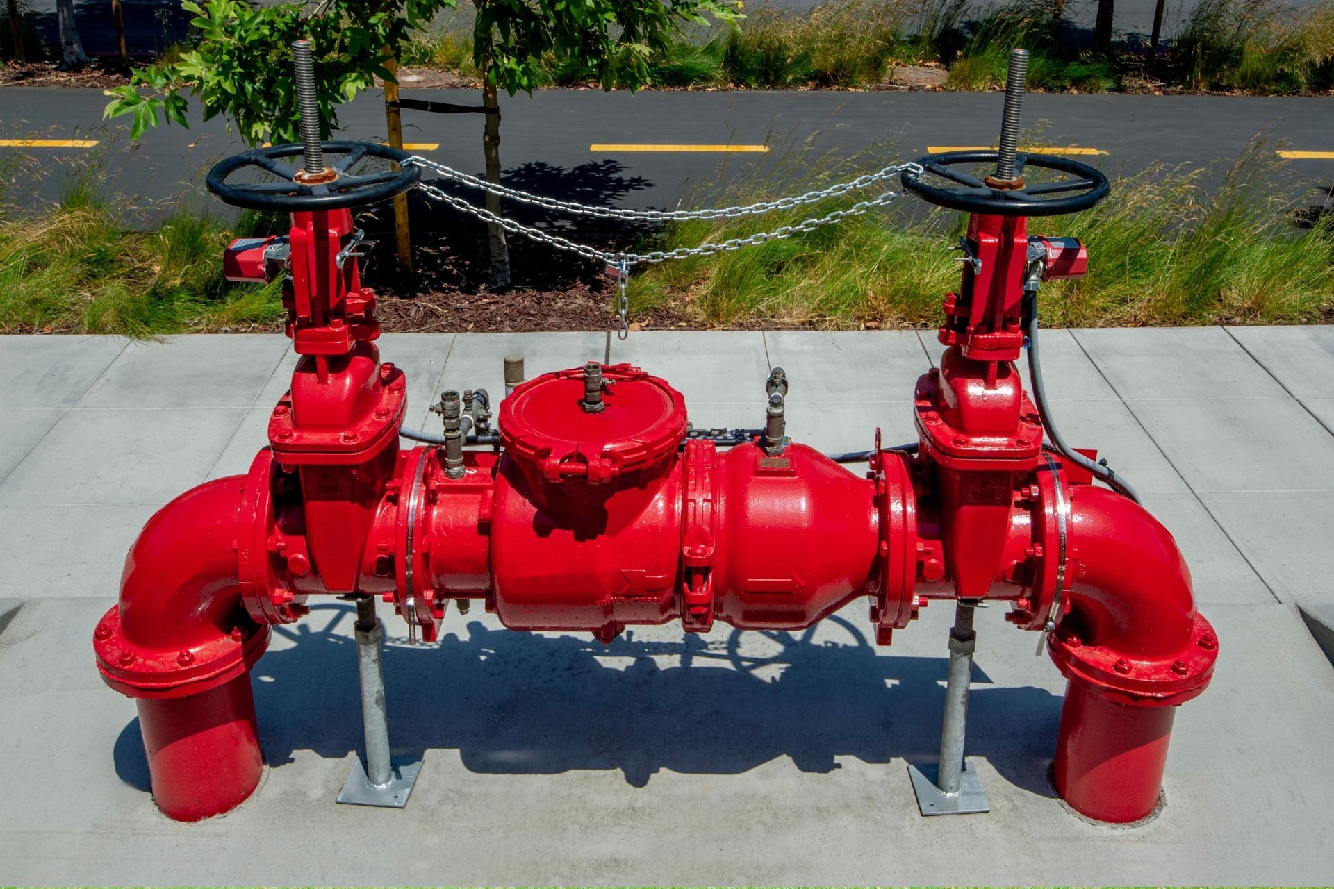 Red industrial fire suppression valve assembly with red pipes and wheel valves beside a sidewalk