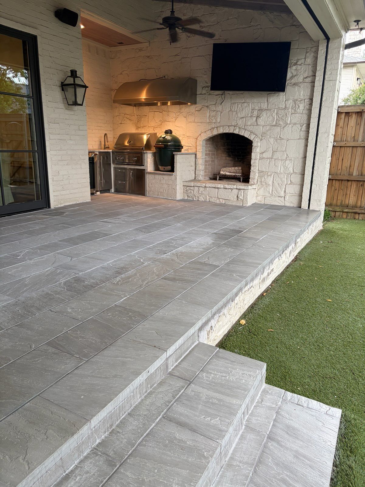 Covered backyard patio with outdoor kitchen, brick fireplace, TV, and gray stone flooring beside artificial turf.