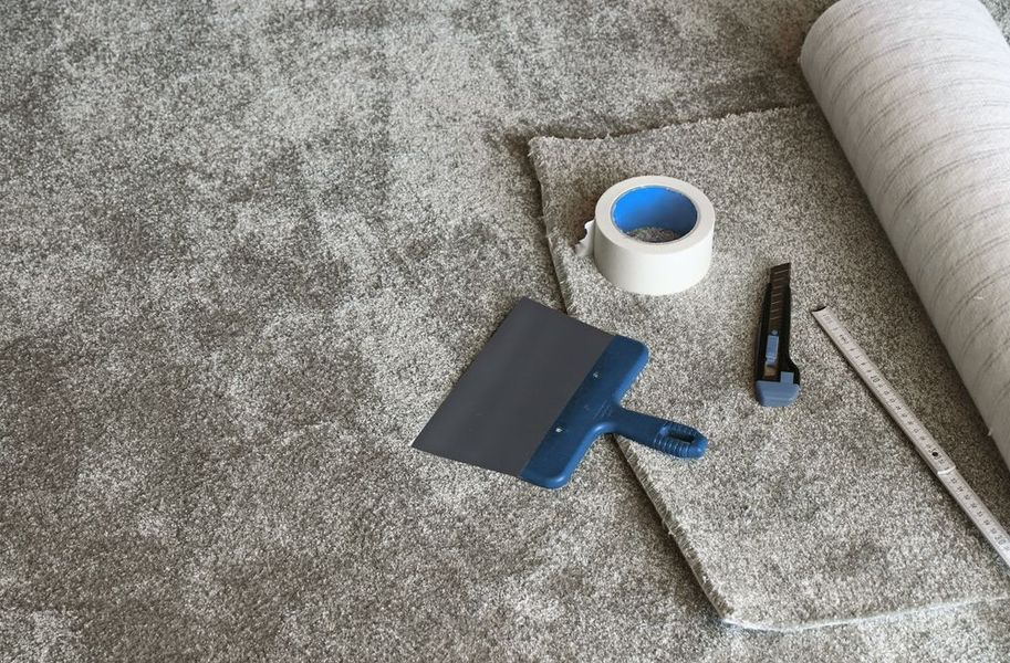 Tools on a gray carpet: blue putty knife, tape roll, utility knife, and metal rod.