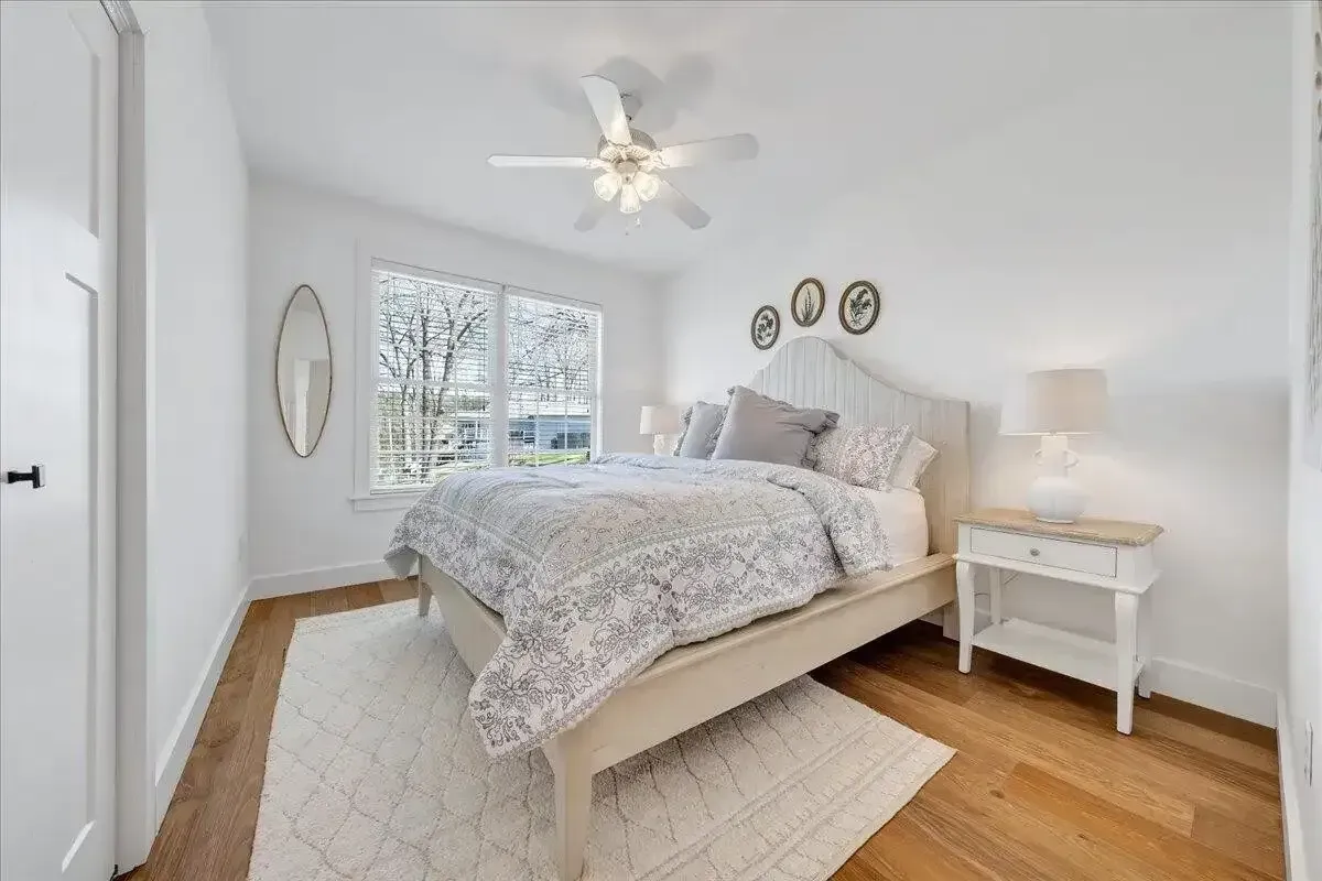 Bright white bedroom with a bed, nightstand, lamp, window, and ceiling fan on hardwood floors.