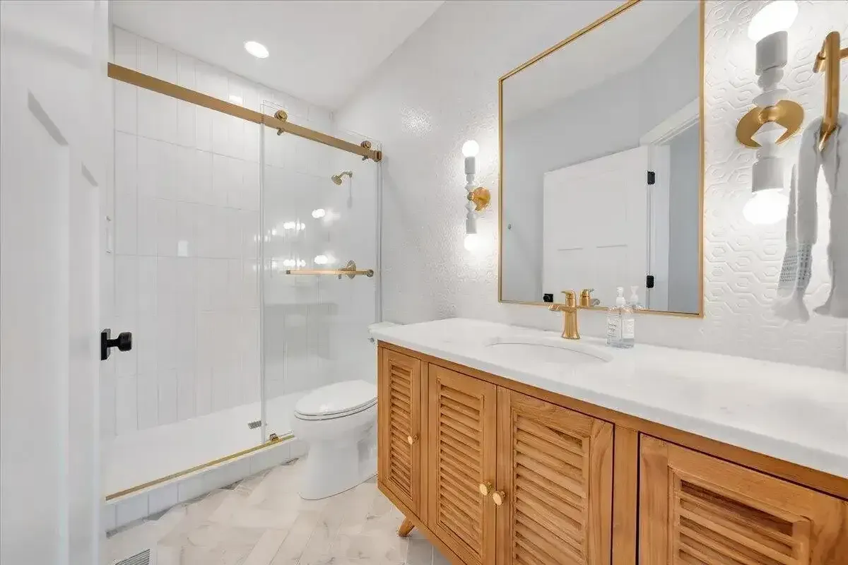 Bright bathroom with glass shower, wood vanity, large mirror, and gold fixtures
