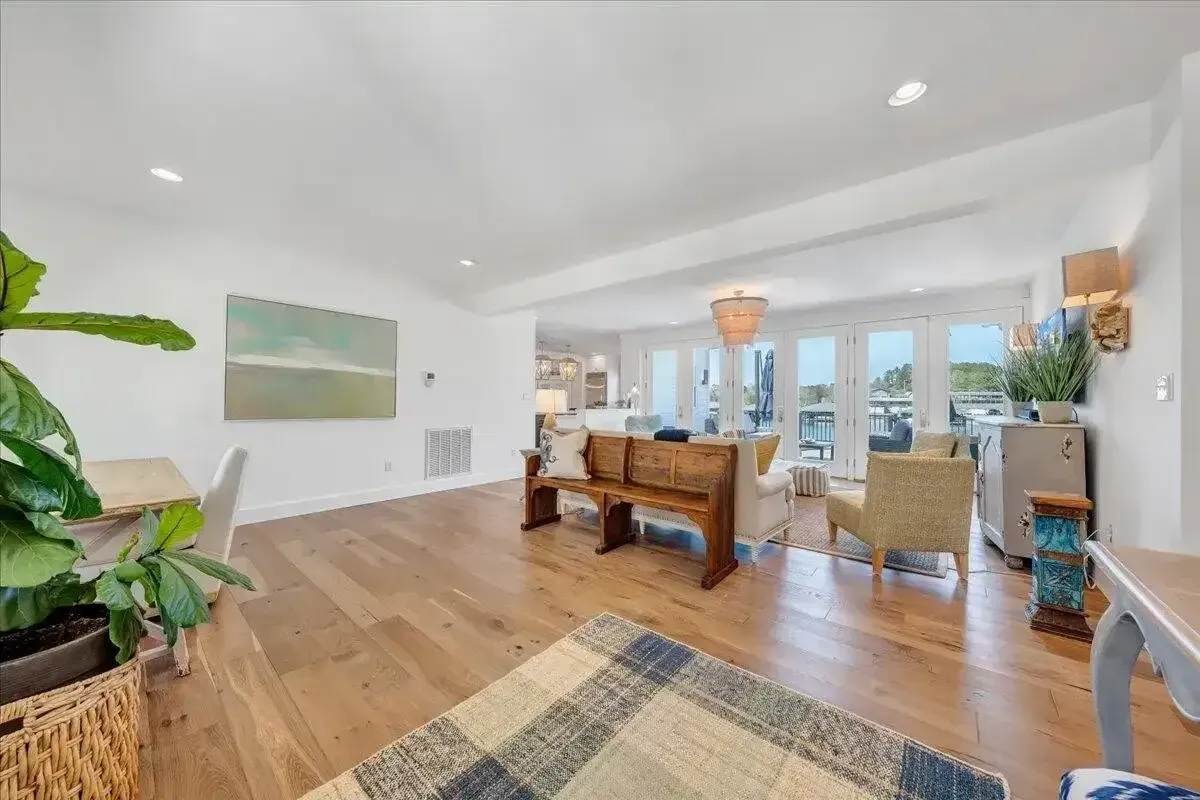 Bright open living room with wood floors, white walls, plants, and a view into the dining area and kitchen.