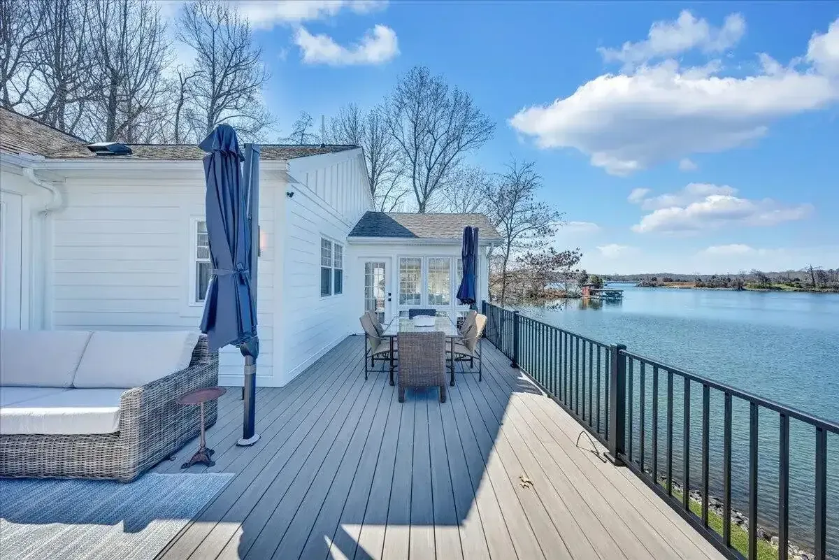 White waterfront deck with tables, railings, and blue umbrellas beside a calm lake under a sunny sky